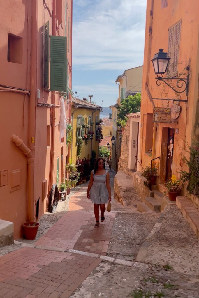A woman strolls down a charming, sunlit alleyway in Menton’s Old Town, surrounded by pastel-colored buildings and glimpses of the sea—one of the most picturesque things to do in Menton, France.