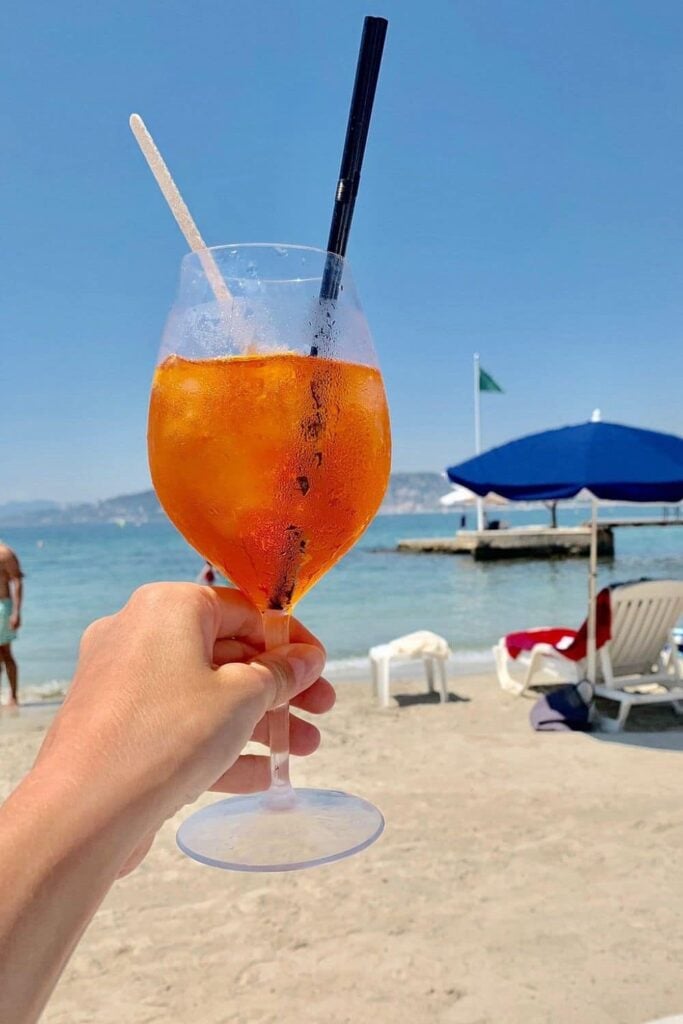 Hand holding an Aperol Spritz cocktail with ice on a sunny day at Belles Rives, a stylish beach club in Antibes, with the sandy beach and blue umbrellas in the background.