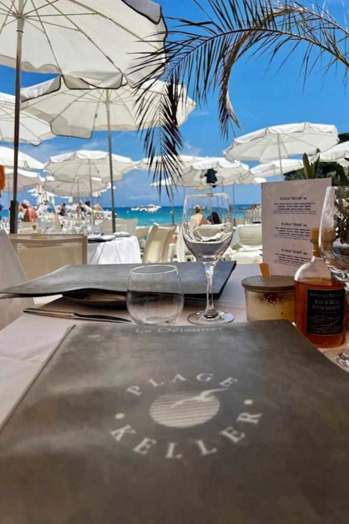 Table set for dining with menus and glasses under white umbrellas at Plage Keller, a renowned beach club in Antibes, with a clear view of the turquoise sea and bright blue sky in the background.