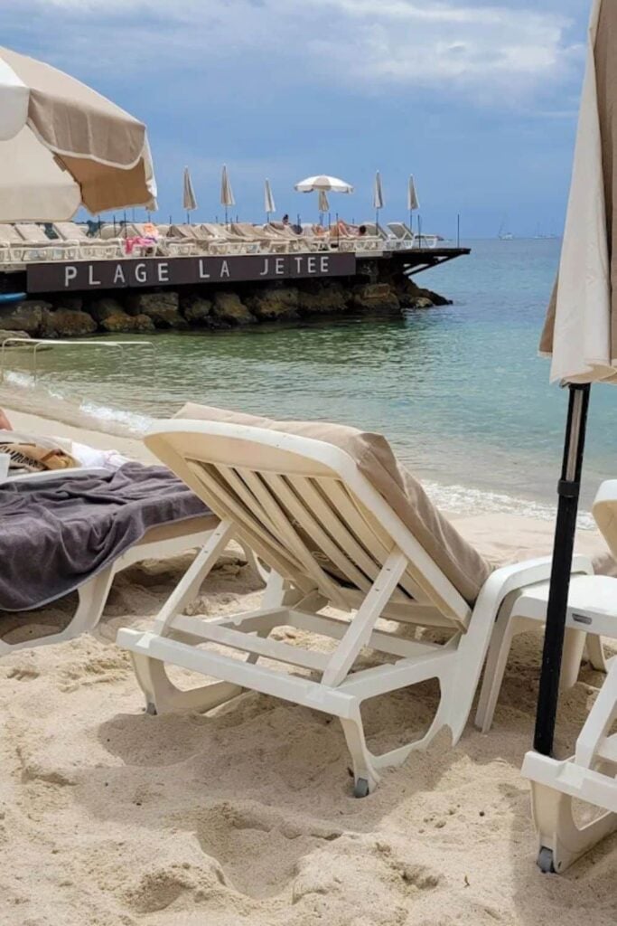 Empty sun loungers and umbrellas on the sandy shore at Plage La Jetée, a charming beach club in Antibes, with calm turquoise waters and cloudy sky in the background.