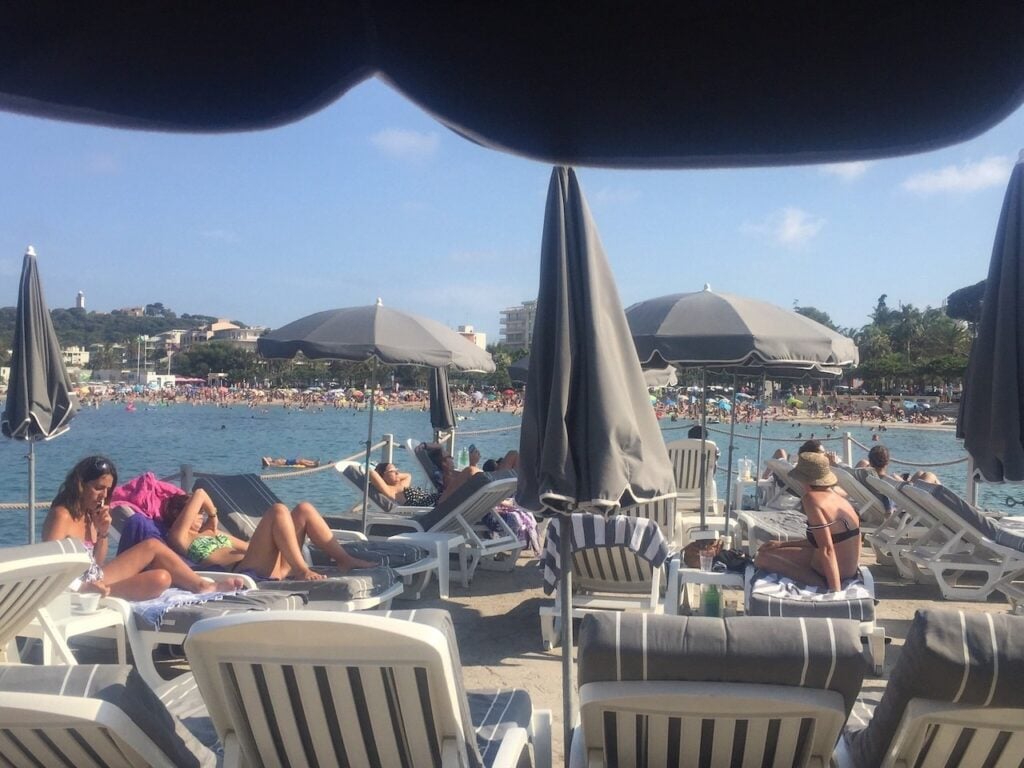 Visitors lounging on white cushioned chairs under gray umbrellas at Royal Beach, one of the elegant beach clubs in Antibes, with the sparkling sea and sandy beach in the background.