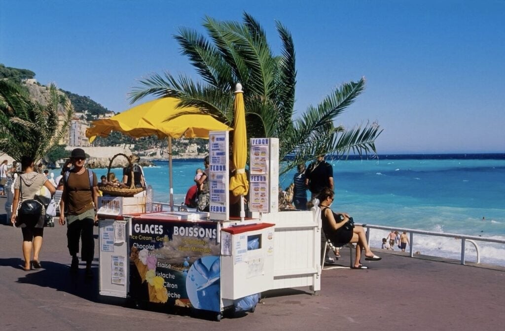 Discover the best ice cream in Nice at a lively beachfront kiosk, shaded by a yellow umbrella and surrounded by palm trees, offering a variety of gelato flavors against the stunning backdrop of the Mediterranean Sea.
