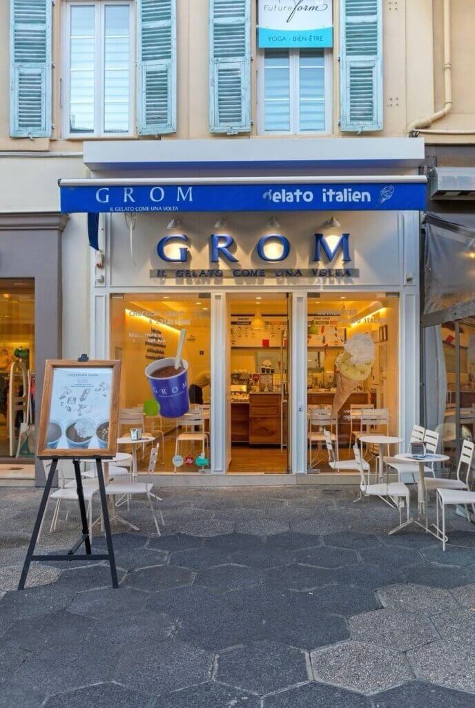 The inviting facade of GROM ice cream shop in Nice, France, with its signature blue and white branding, offers a glimpse into the classic Italian gelateria experience, complete with outdoor seating and a menu board showcasing artisanal flavors.