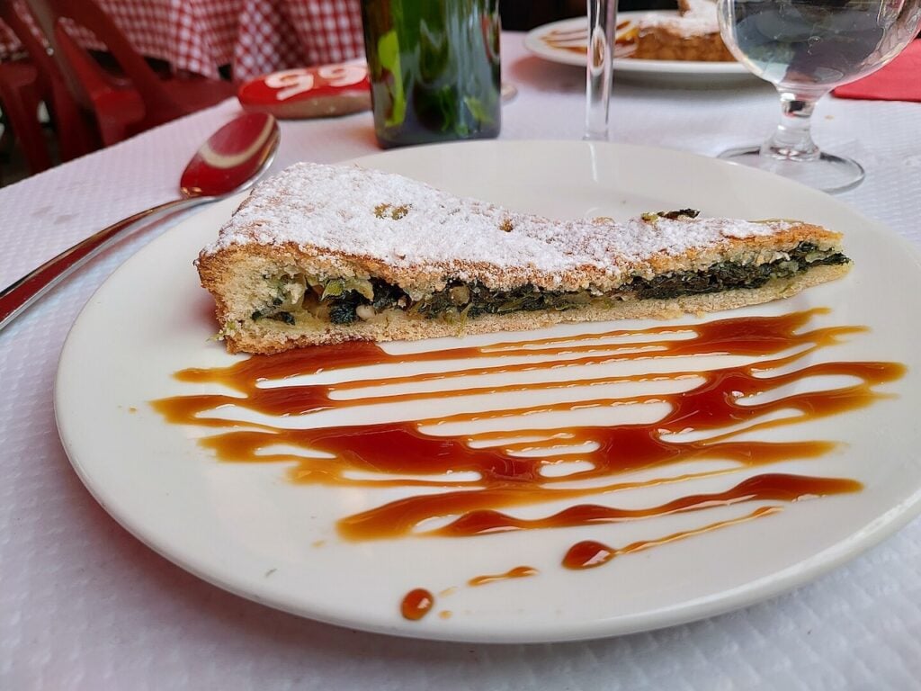 A slice of Cuisine Niçoise traditional tourte de blettes, a Swiss chard and pine nut tart, dusted with powdered sugar and served with a caramel drizzle.