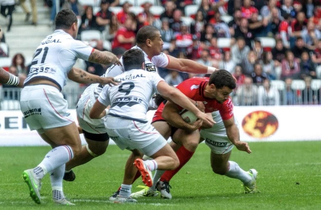 Things to do in Toulon: Action shot from a rugby match featuring RC Toulon in Toulon, France. A player in red is in possession of the ball and is being tackled by opposing players in white. The intensity of the game is evident in the players' focused expressions and dynamic poses. The stadium is partially visible in the background with spectators looking on, adding to the sense of excitement and competition of the live sporting event.