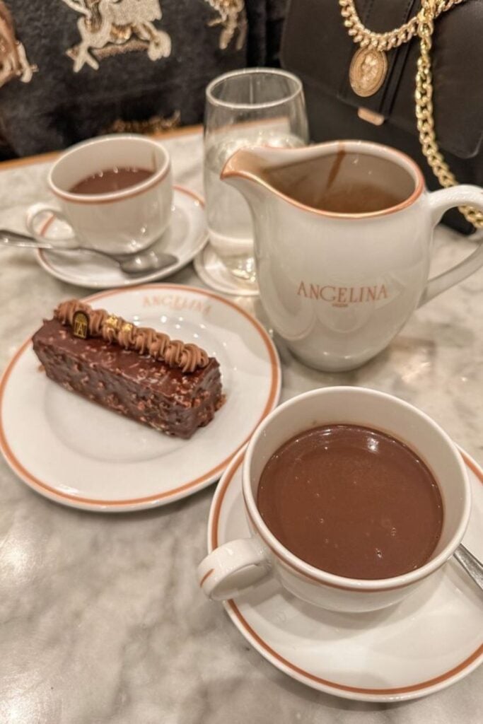 A table at Angelina in Paris, known for the best hot chocolate in Paris, featuring two cups of the signature drink, a pitcher, and a chocolate dessert on elegant white china.