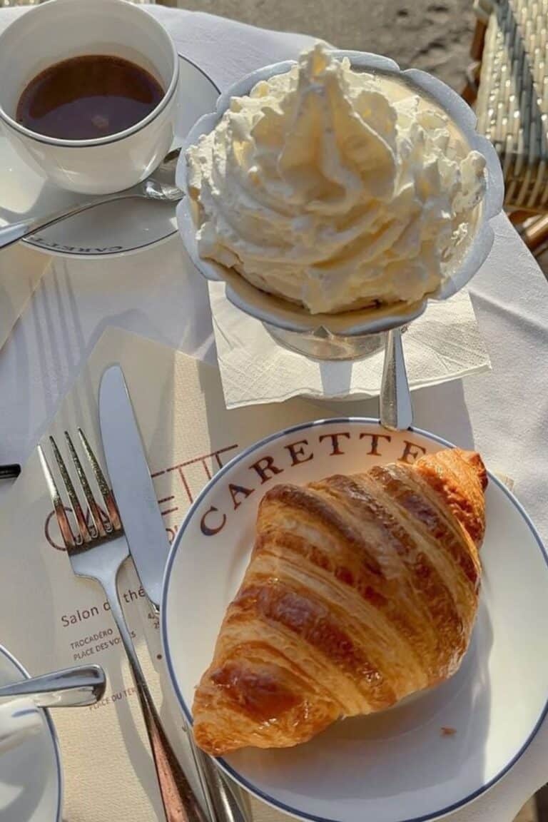 A sunlit table at Carette in Paris showcasing the best hot chocolate in Paris, served with a mountain of whipped cream and a flaky, golden croissant.