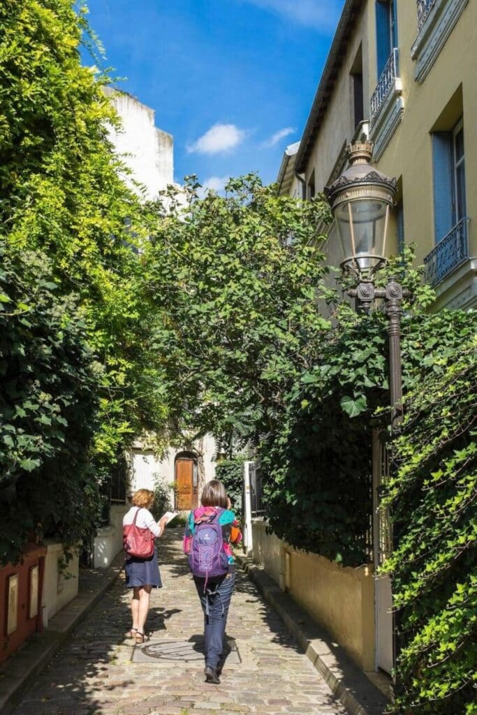 Two women walking along the cobblestone path of Square des Peupliers, a leafy and quiet residential lane, one of the hidden gems in Paris known for its village-like atmosphere and greenery.