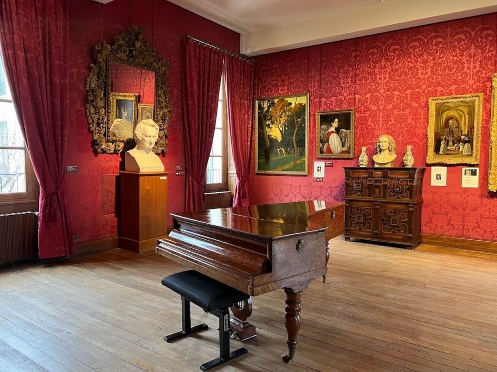 A room in the Maison de Victor Hugo in Paris, richly decorated with red damask wallpaper and heavy drapery. A grand piano sits at the center of the wooden floor, while framed artworks and busts of Hugo adorn the walls and surfaces, reflecting the literary giant’s grandeur and the 19th-century aesthetic.