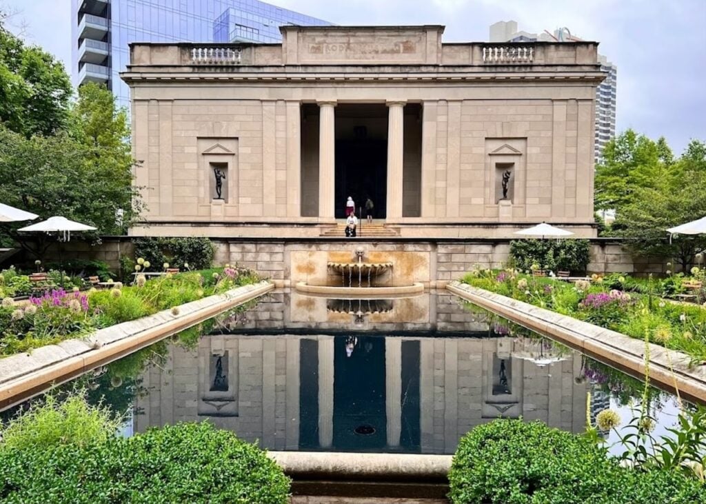 The tranquil garden of the Rodin Museum in Paris, featuring a long rectangular reflecting pool leading to the elegant neoclassical façade of the museum. Lush flowerbeds and well-manicured hedges line the pool, and sculptures are strategically placed, offering a serene outdoor gallery experience to visitors.