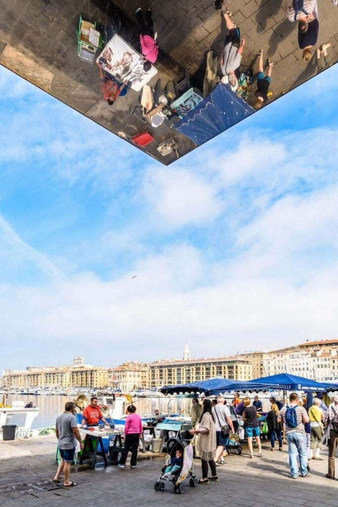 A bustling fish market at Marseille's Old Port, reflected in the mirrored canopy, where visitors can soak in the atmosphere and find unique Marseille souvenirs.