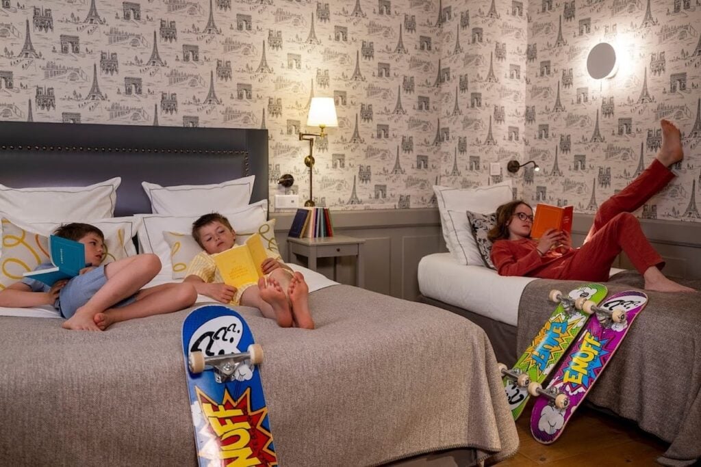 Three kids relaxing and reading books on beds in a cozy room at Paris Hotels with Connecting Rooms, decorated with Eiffel Tower wallpaper and skateboards nearby.