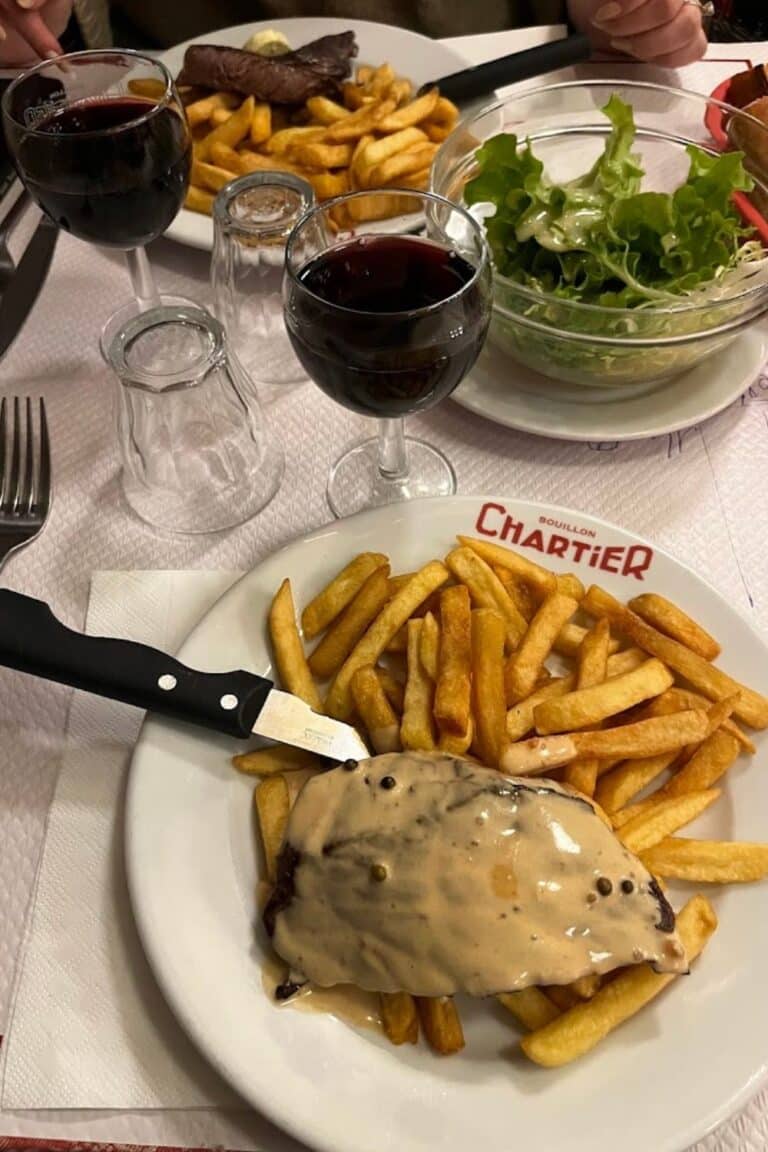 Peppercorn steak with fries served at Bouillon Chartier, one of the most iconic bouillon restaurants in Paris, alongside glasses of red wine and a simple green salad.