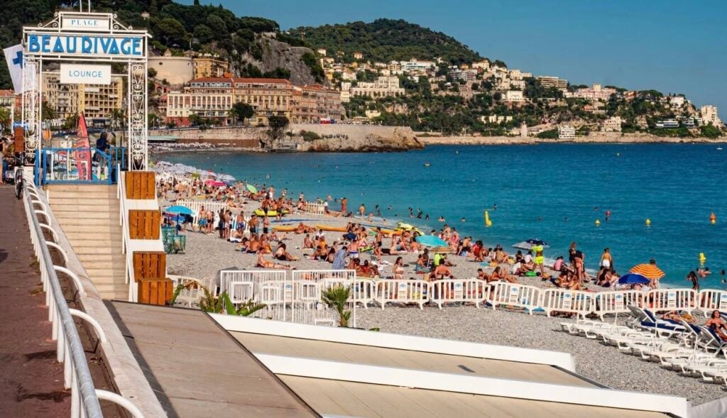 Busy pebble beach at Beau Rivage Lounge, one of the lively beach clubs in Nice with sunbathers enjoying the Mediterranean Sea and hillside buildings in the background.