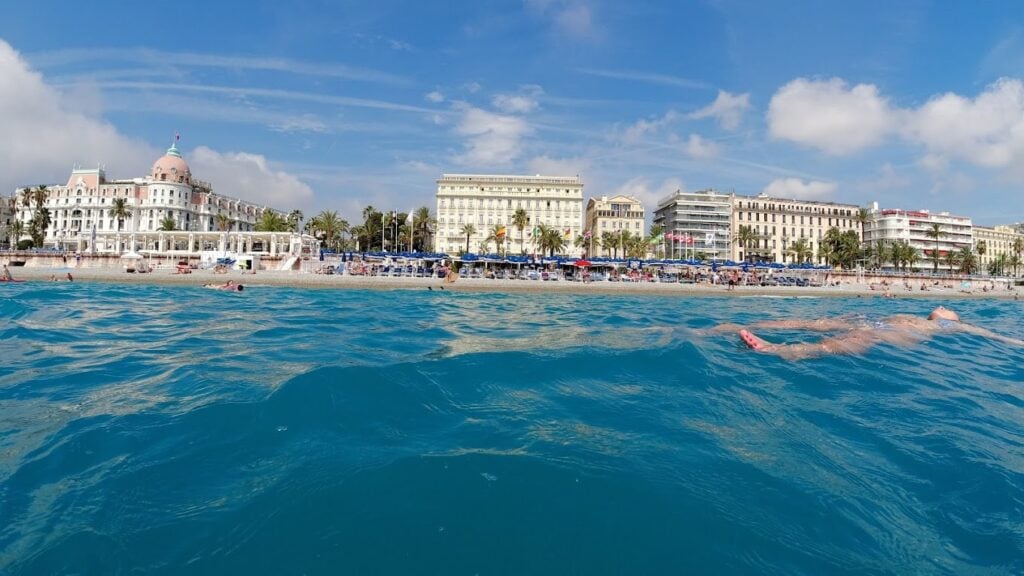 View from the water of Blue Beach and other beach clubs in Nice along the Promenade des Anglais, showing swimmers in the Mediterranean Sea and iconic seaside hotels in the background.