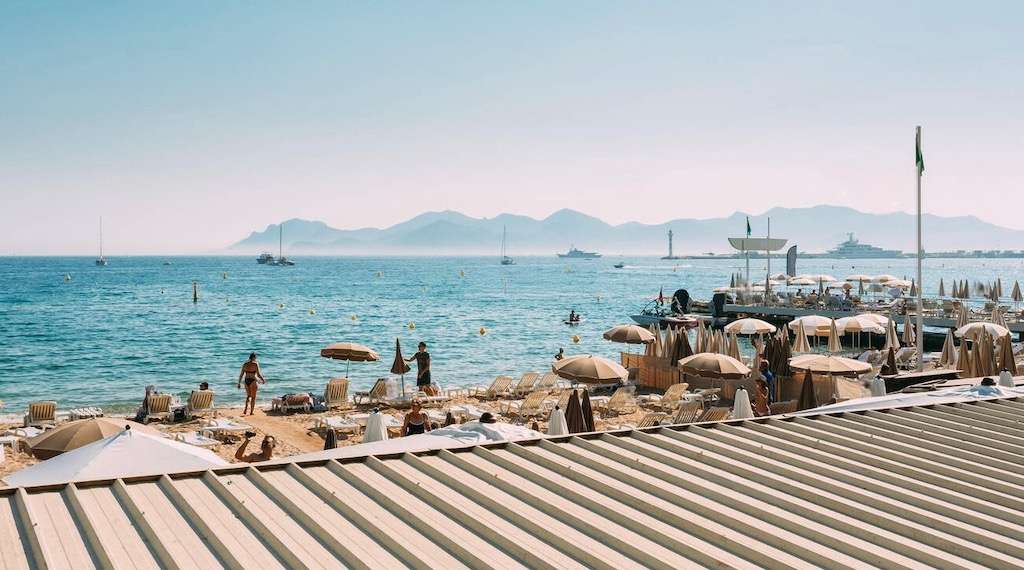 Sunny beach scene at Cannes France showing sandy shore, sun loungers, umbrellas, and people enjoying the water at famous Cannes beach clubs, with mountains in the distance across the Mediterranean.