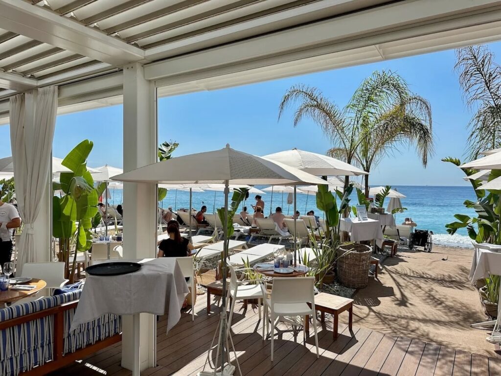Shaded outdoor dining area overlooking sun loungers and white umbrellas at Le Galet, one of the chic beach clubs in Nice with clear blue skies and Mediterranean sea in the background.