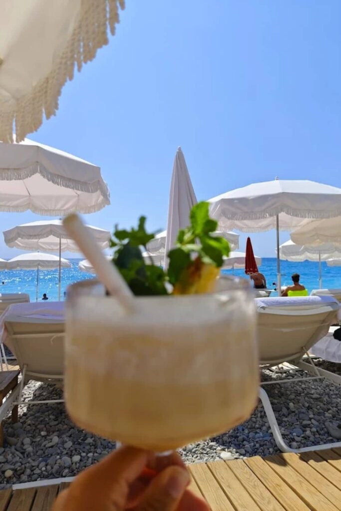 Close-up of a refreshing cocktail held up against the backdrop of white umbrellas and sun loungers at Le Negresco, a timeless and casual beach club in Nice France.
