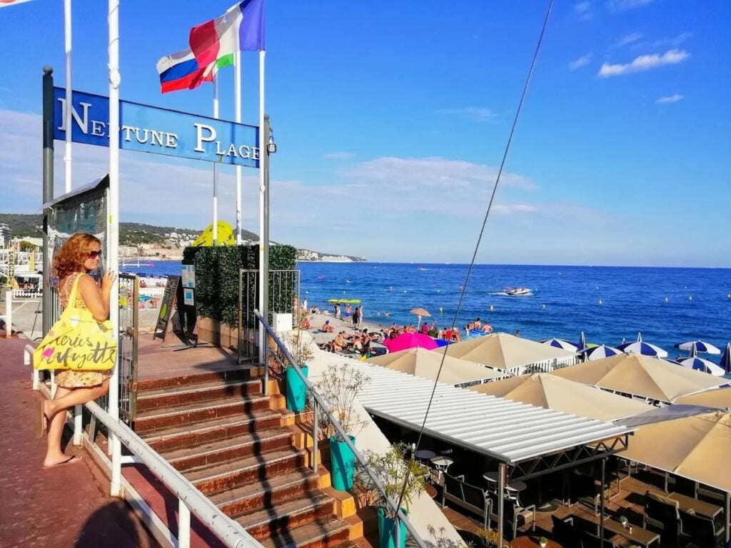 Woman standing near the entrance to Neptune Plage, with beige umbrellas and sun loungers lining the beach at one of the favorite beach clubs in Nice France, against a bright blue sky and Mediterranean Sea.