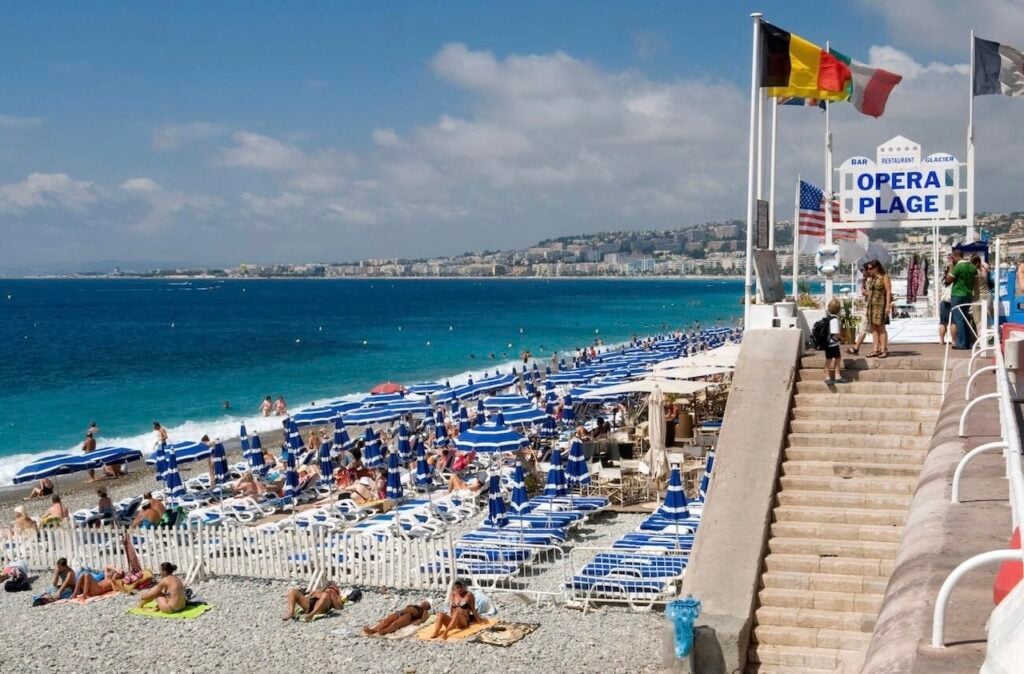 View of Opera Plage’s private beach with rows of blue umbrellas and sun loungers along the pebble shore at one of the popular beach clubs in Nice, France.