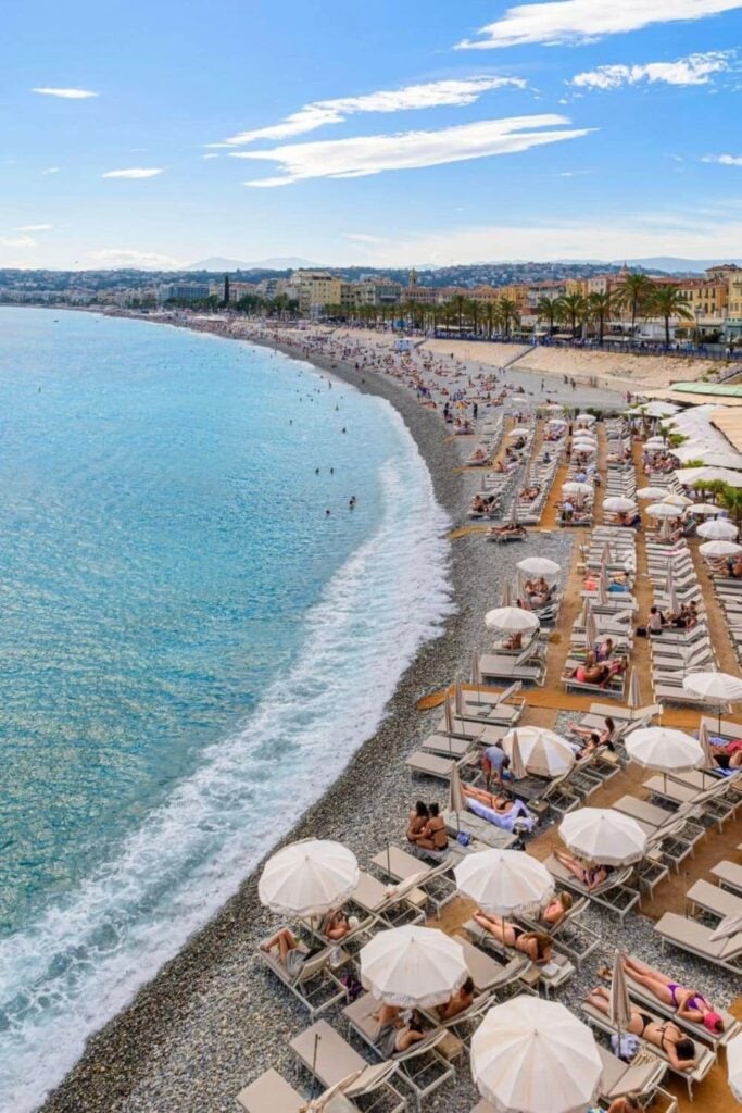A sunny view of a busy private beach along the Promenade des Anglais in Nice, with rows of lounge chairs and white umbrellas at one of the popular beach clubs in Nice France.