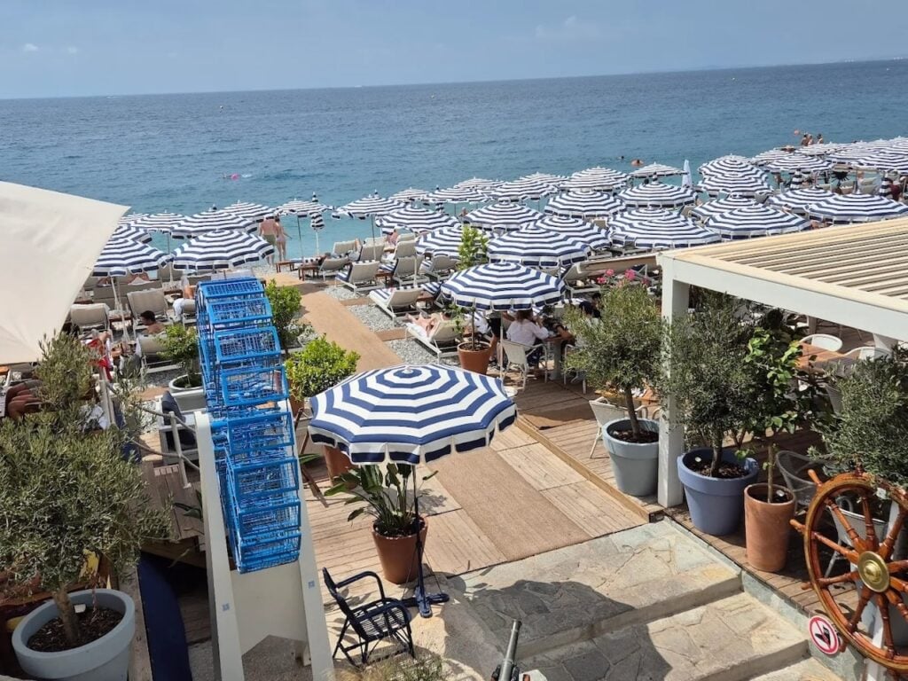 Rows of blue and white striped umbrellas and sun loungers line the pebble beach at Ruhl Plage, one of the iconic beach clubs in Nice with the Mediterranean Sea stretching into the horizon.
