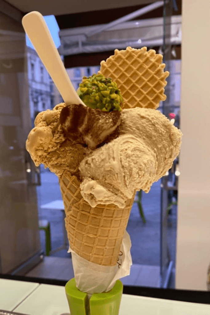 Indulge in the best ice cream in Nice with this towering waffle cone, generously scooped with rich, creamy gelato, topped with a crunchy wafer and a sprinkle of vibrant green pistachios, served with a spoon, ready to savor.