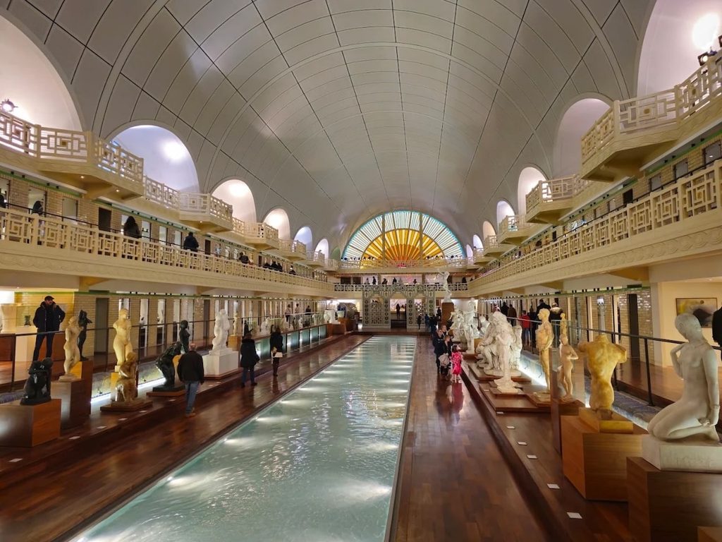 Interior of La Piscine Museum in Roubaix, featuring sculptures displayed around the long former Art Deco swimming pool beneath a high arched glass ceiling and colorful sunburst stained-glass window.