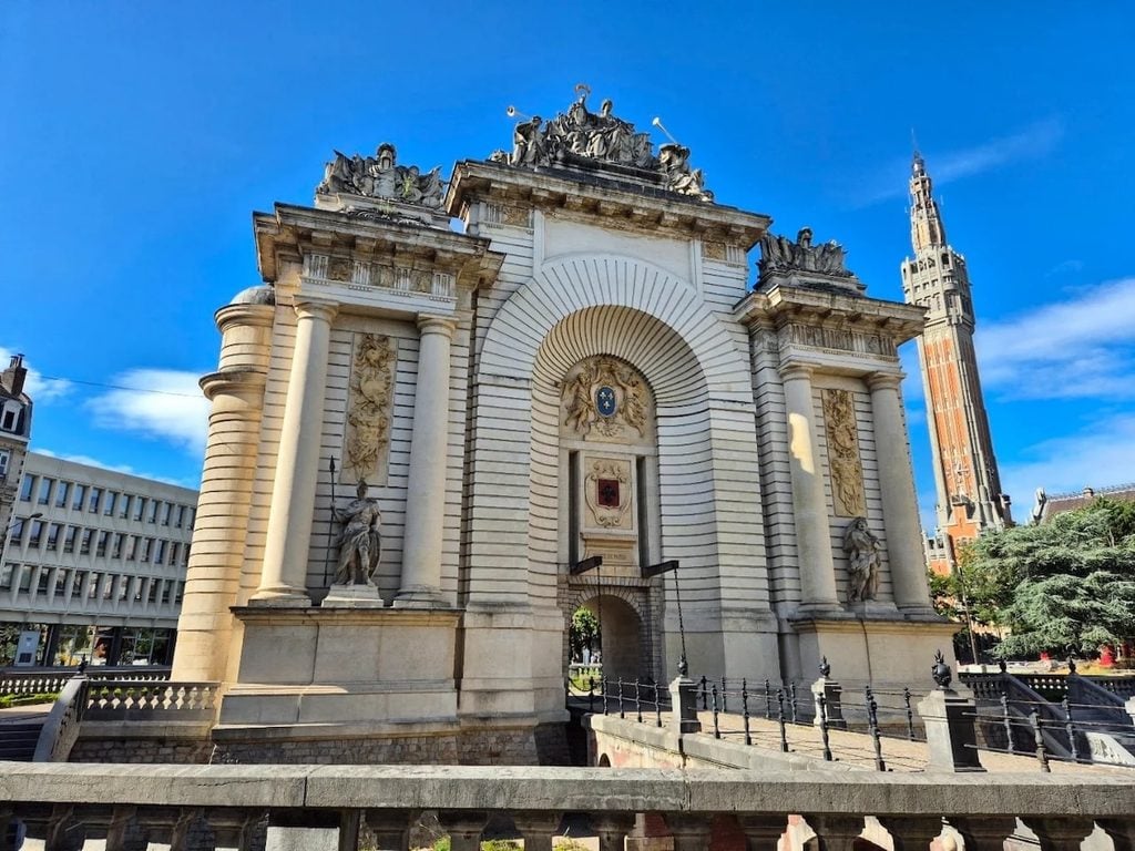 The ornate stone Porte de Paris triumphal arch in Lille, featuring a large central passage, decorative columns, sculptures, and a crowned figure at the top, surrounded by green trees beneath a blue sky.
