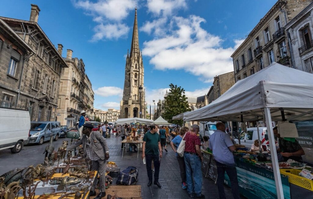 Shoppers browse through a bustling brocante market on the streets of Bordeaux in the Winter, with the historic spire of Saint-Michel standing tall against a clear sky.