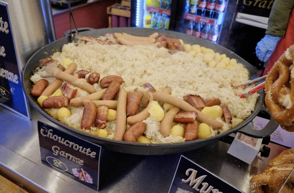 A giant pan filled with traditional Alsatian choucroute garnie, featuring sauerkraut, various sausages, and potatoes, ready to be savored at the Lille Christmas Market, under a sign that reads 'Choucroute Garnie 8€'.