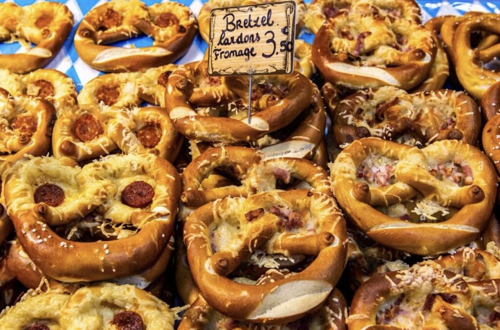 Freshly baked pretzels topped with melted cheese and diced ham on display at Lille Christmas market, with a sign indicating 'Bretzel Lardon Fromage' for 3.5 euros, capturing the delicious local street food offerings.