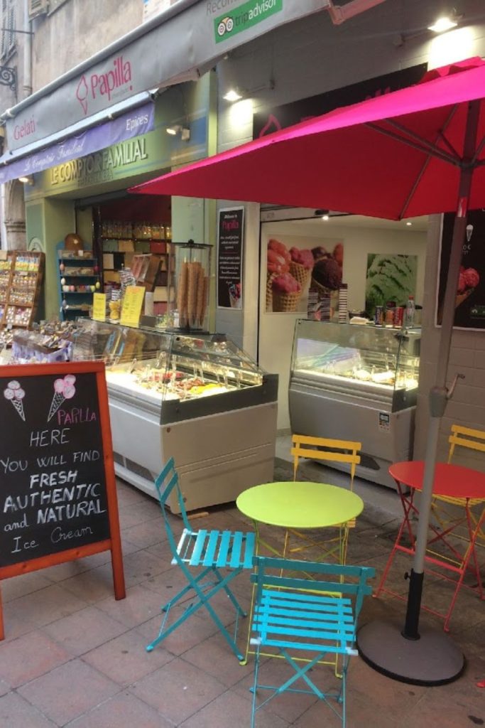 Colorful street view of Glacier Papilla, an ice cream shop in Nice, with a chalkboard sign stating 'Here you will find FRESH AUTHENTIC and NATURAL Ice Cream'. Brightly colored outdoor chairs and a vivid pink umbrella invite visitors to enjoy their gelato treats.