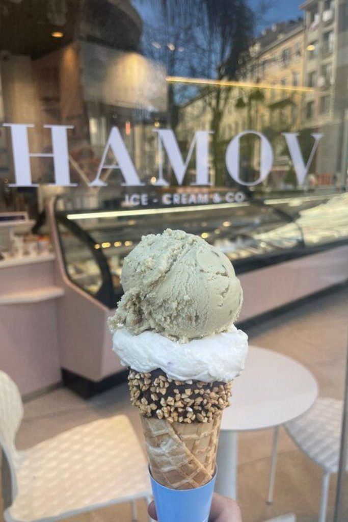 Savor the flavors of the best ice cream in Nice with a close-up of a hand holding a cone topped with a creamy scoop encrusted with crunchy nuts, in front of the HAMOV Ice Cream & Co shop window, reflecting the charming city streets.
