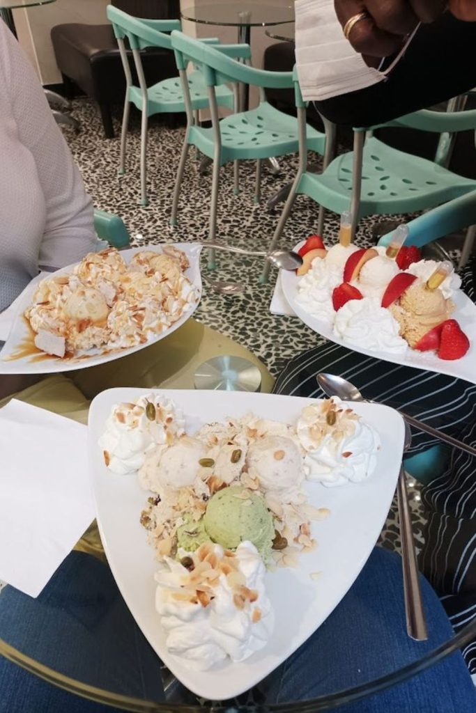 A heart-shaped dish filled with assorted scoops of ice cream topped with whipped cream, sliced almonds, and pumpkin seeds, shared by two people at a cafe with mint green chairs in the background, capturing the essence of a sweet treat in Nice.