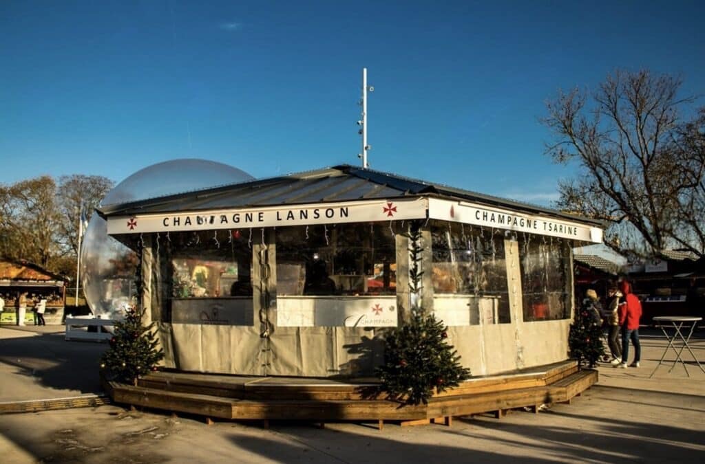 A cozy outdoor champagne stall at the Reims Christmas Market, adorned with festive decorations and a clear dome structure on a sunny day.