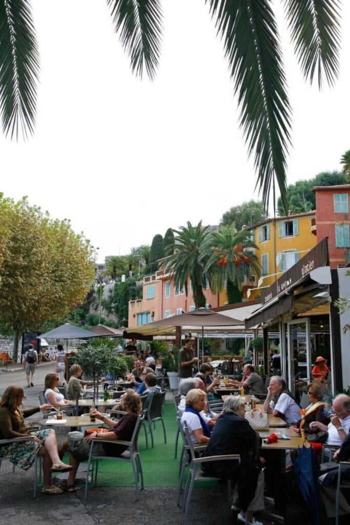Outdoor café scene with people dining under palm trees in one of the charming French Riviera cities, featuring colorful buildings and a relaxed Mediterranean atmosphere.