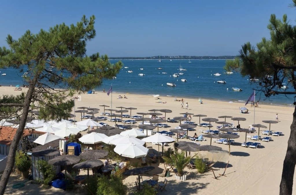 The serene Arcachon beach near Bordeaux in the Winter, with its sandy shores lightly populated by beachgoers, framed by green pines against a backdrop of calm blue waters and moored boats.