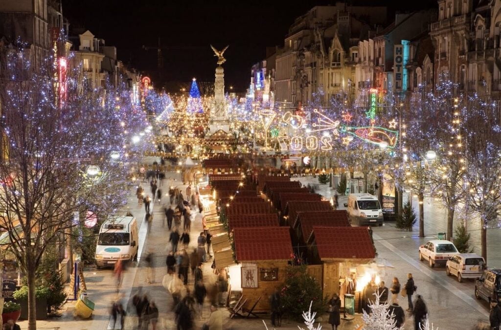 A magical evening view of the Reims Christmas Market, bustling with people amid rows of market stalls, surrounded by trees and street lights festooned with twinkling blue and white Christmas lights, creating a winter wonderland atmosphere.