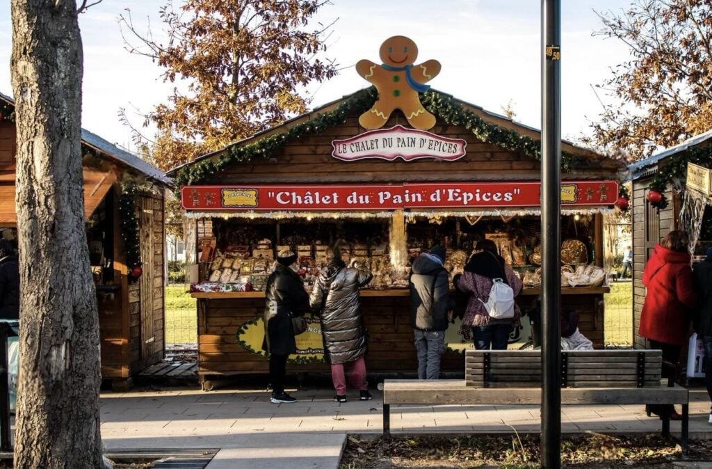 Shoppers gather at 'Chalet du Pain d'Épices' at the Reims Christmas Market, featuring a large gingerbread man decoration on top and a variety of gingerbread treats on display, offering a traditional taste of festive sweets.