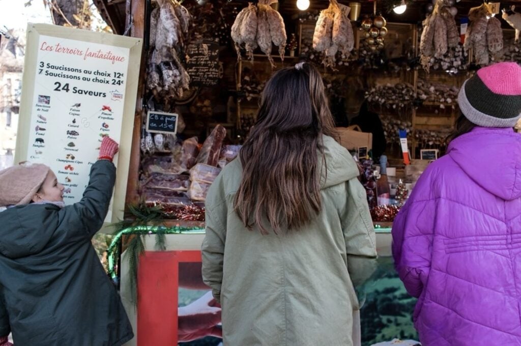 Patrons at the Reims Christmas Market peruse a selection of truffled sausages, with a menu board listing '24 Saveurs' of handcrafted charcuterie, capturing the market's rich culinary offerings.