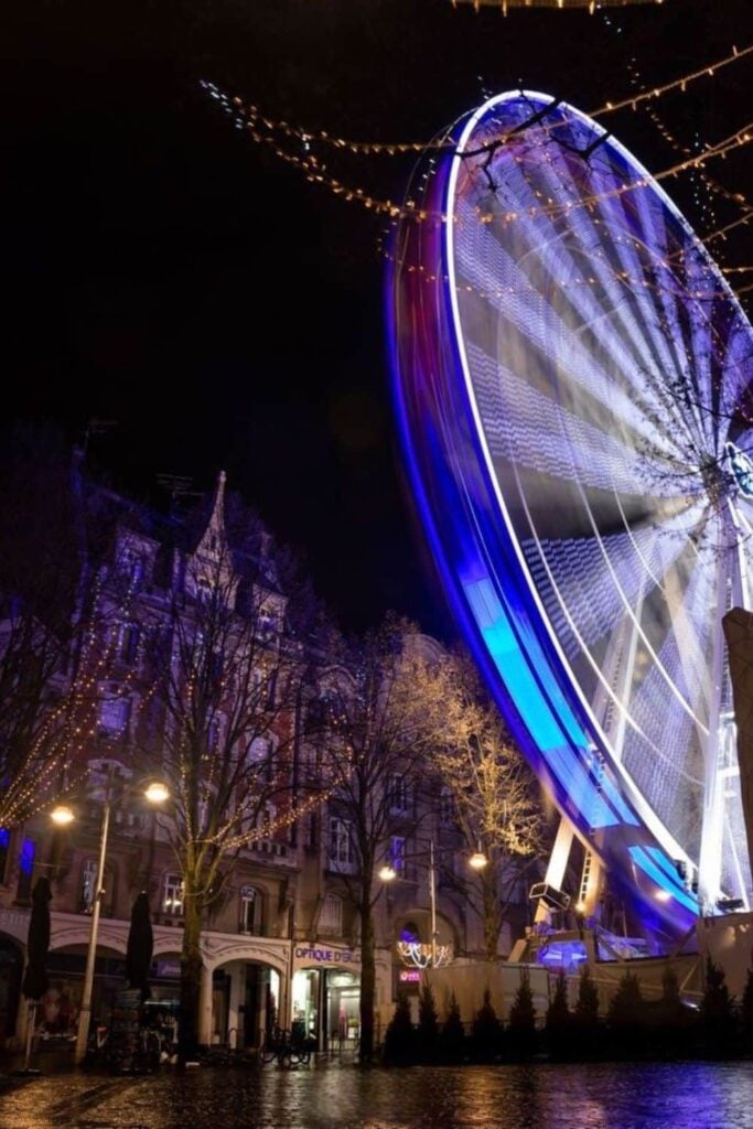 A Ferris wheel captured in motion with a long exposure, creating a blur of lights against the night sky at the Reims Christmas Market, with surrounding trees and festive decorations enhancing the holiday spirit.