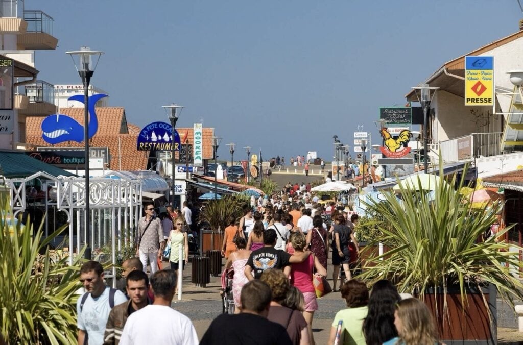 day trips from Bordeaux France: A bustling seaside promenade in Biscarrosse, France, teeming with tourists exploring various shops and eateries under a bright blue sky, with colorful signs and palm plants adding to the vibrant summer atmosphere.