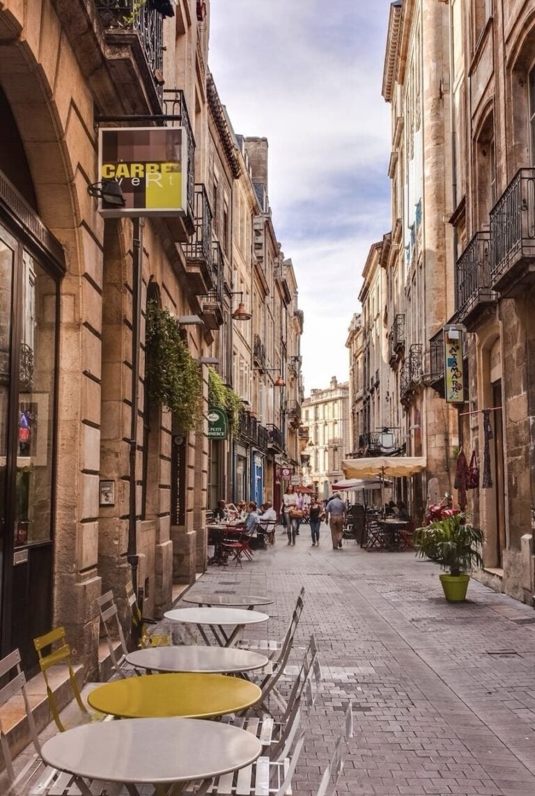 A quaint street in Bordeaux, lined with traditional limestone buildings and inviting café terraces. The picture captures the relaxed ambiance of city life, with people dining and strolling, and vibrant shop signs adding a pop of color to the historic charm of the pedestrian-friendly cobblestone path.