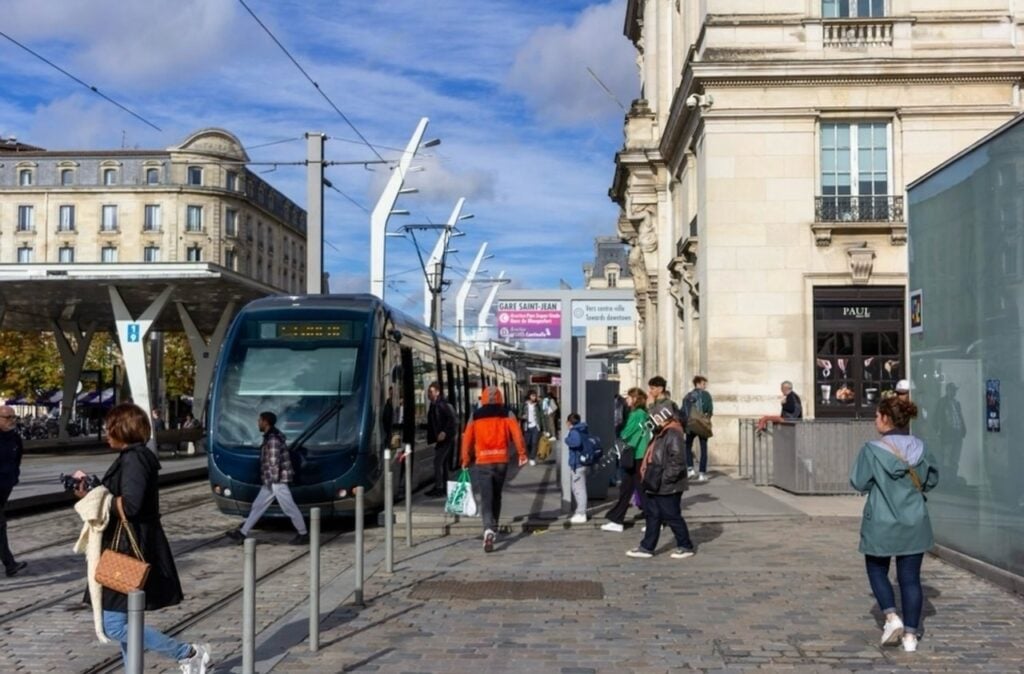 Pedestrians and commuters navigate a tram station in a vibrant urban setting, with a sleek blue tram halted at the stop. The backdrop features classic architecture and clear skies, showcasing a seamless blend of modern transportation and historic city design.