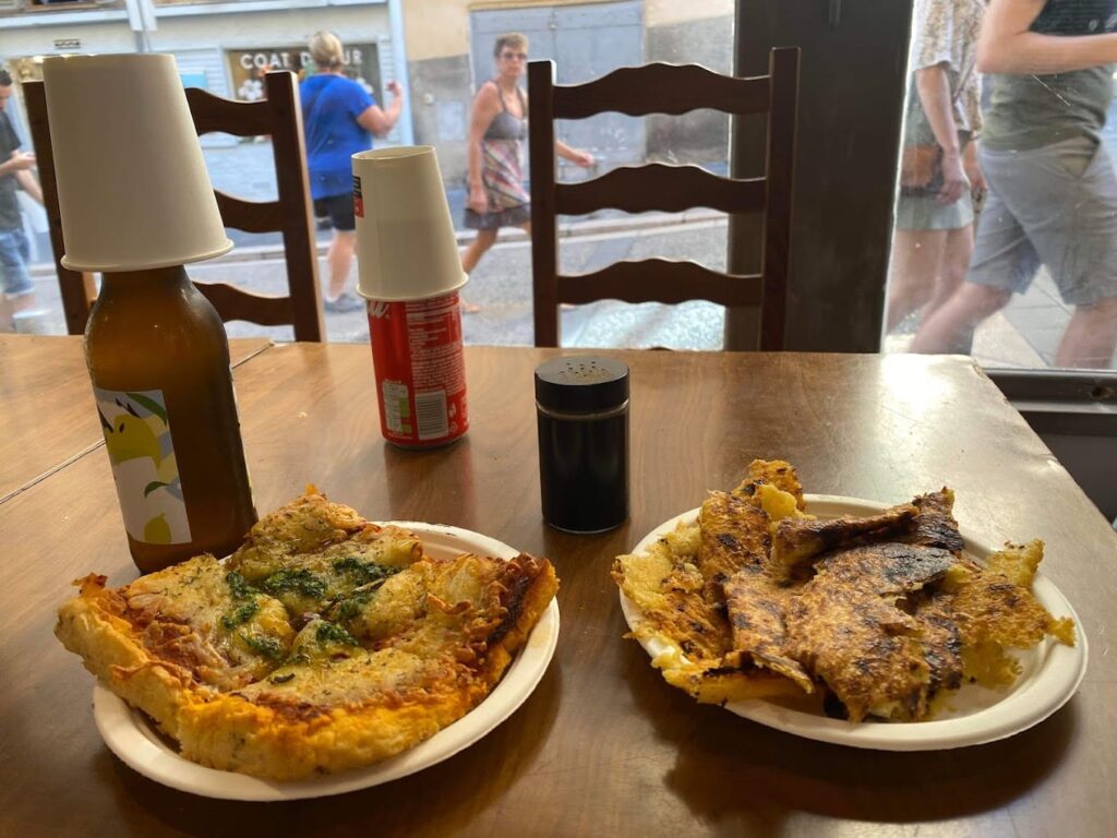 Two plates of traditional Niçoise cuisine, one featuring a slice of pissaladière and another with socca, placed on a wooden table inside a restaurant. Condiment bottles with paper covers and a window offering a view to pedestrians on the street add to the casual dining atmosphere.