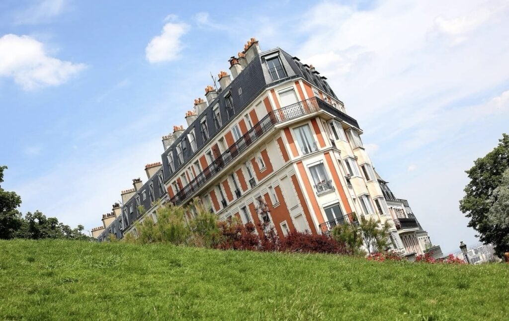 A picturesque 'sinking' house illusion in Montmartre, Paris, with the building appearing tilted due to the sloped grassy foreground, against a clear blue sky.