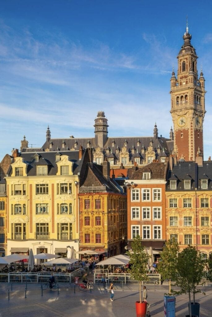 Historic town square under a soft evening light, featuring a towering belfry, and classic European architecture with colorful facades, as people enjoy the outdoor seating of a cafe.