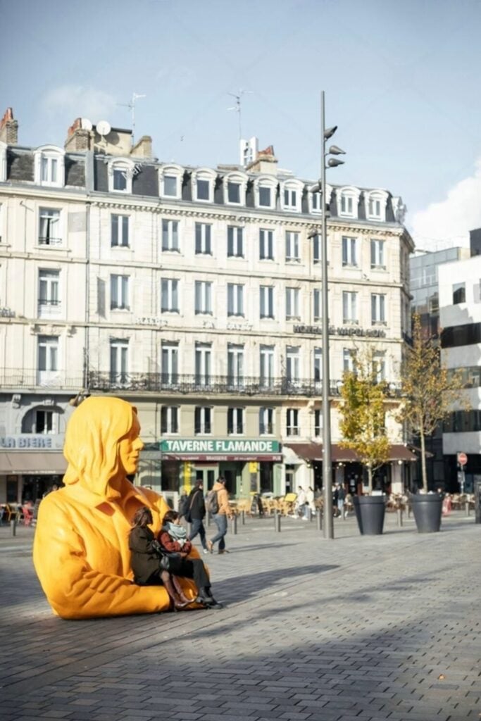 A vibrant yellow sculpture serves as seating in a lively square near historic buildings and cafés, offering a unique spot for relaxation among the many things to do in Lille.