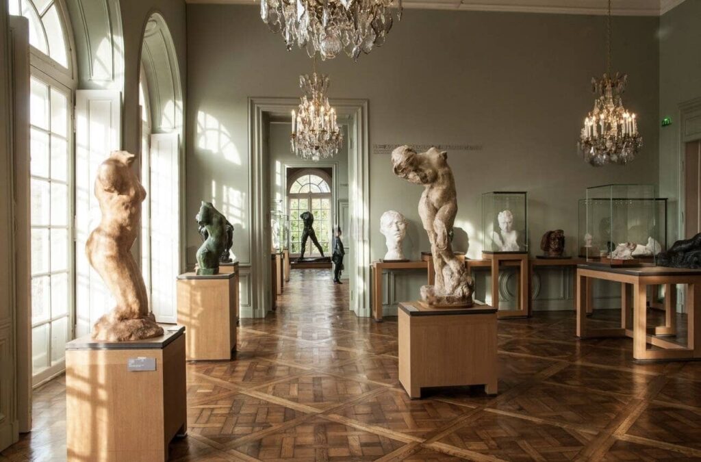 Sunlit interior of a classic sculpture gallery with parquet flooring, featuring an array of marble statues on pedestals and busts within glass cases, complemented by elegant chandeliers and tall arched windows.